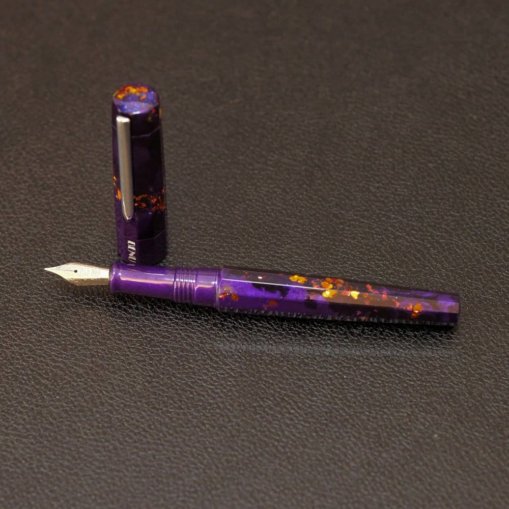 The BENU Euphoria, a faceted fountain pen with 10 sides, and a dark purple acrylic body shot through with small, dark blue glitter and chunky copper glitter, shown against a black, textured background, uncapped