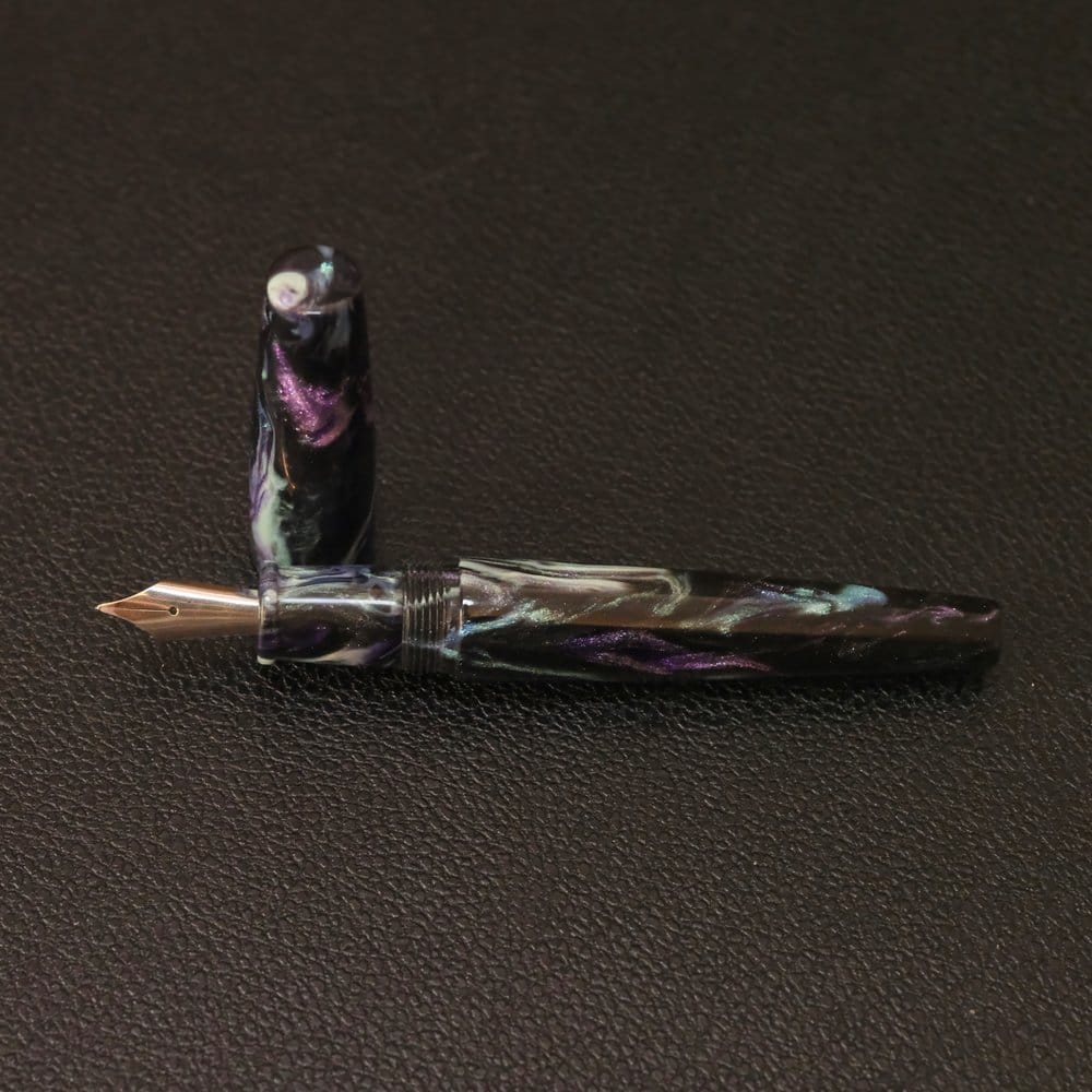 Cigar-shaped fountain pen with flat ends by Skogsy Pens, the Cholla model, in a black acrylic body with pearl, aqua, and purple shimmer swirled throughout, shown on a black textured background, uncapped