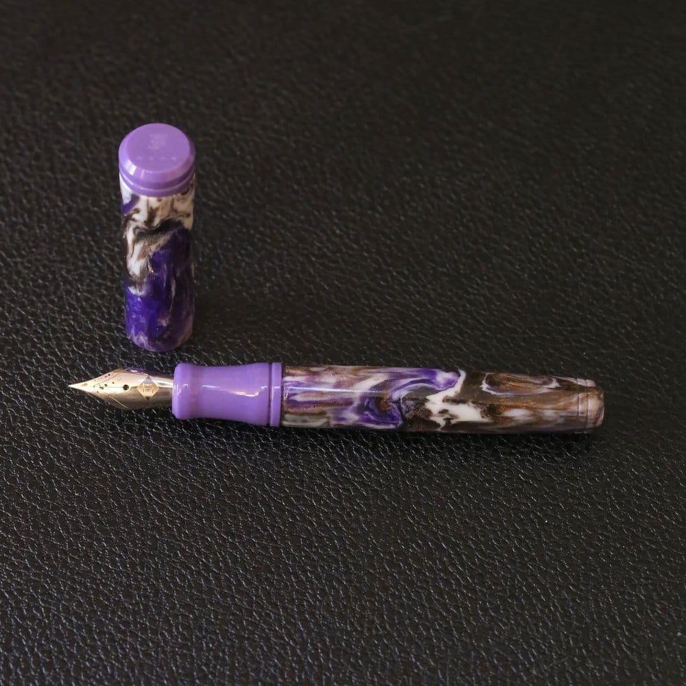 "Pocket-sized Franklin-Christoph fountain pen with medium purple cap finial and grip, and a swirly acrylic body of purple, black, cream, and brown shimmer, shown against a black, textured background, uncapped