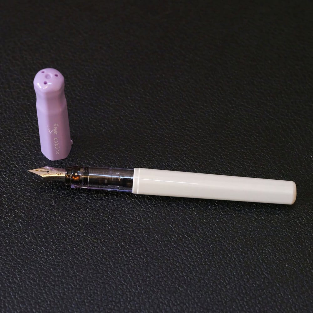 Pilot Kakuno, a plastic fountain pen with a white body, clear grip, and light purple cap, shown against a black, textured background, uncapped