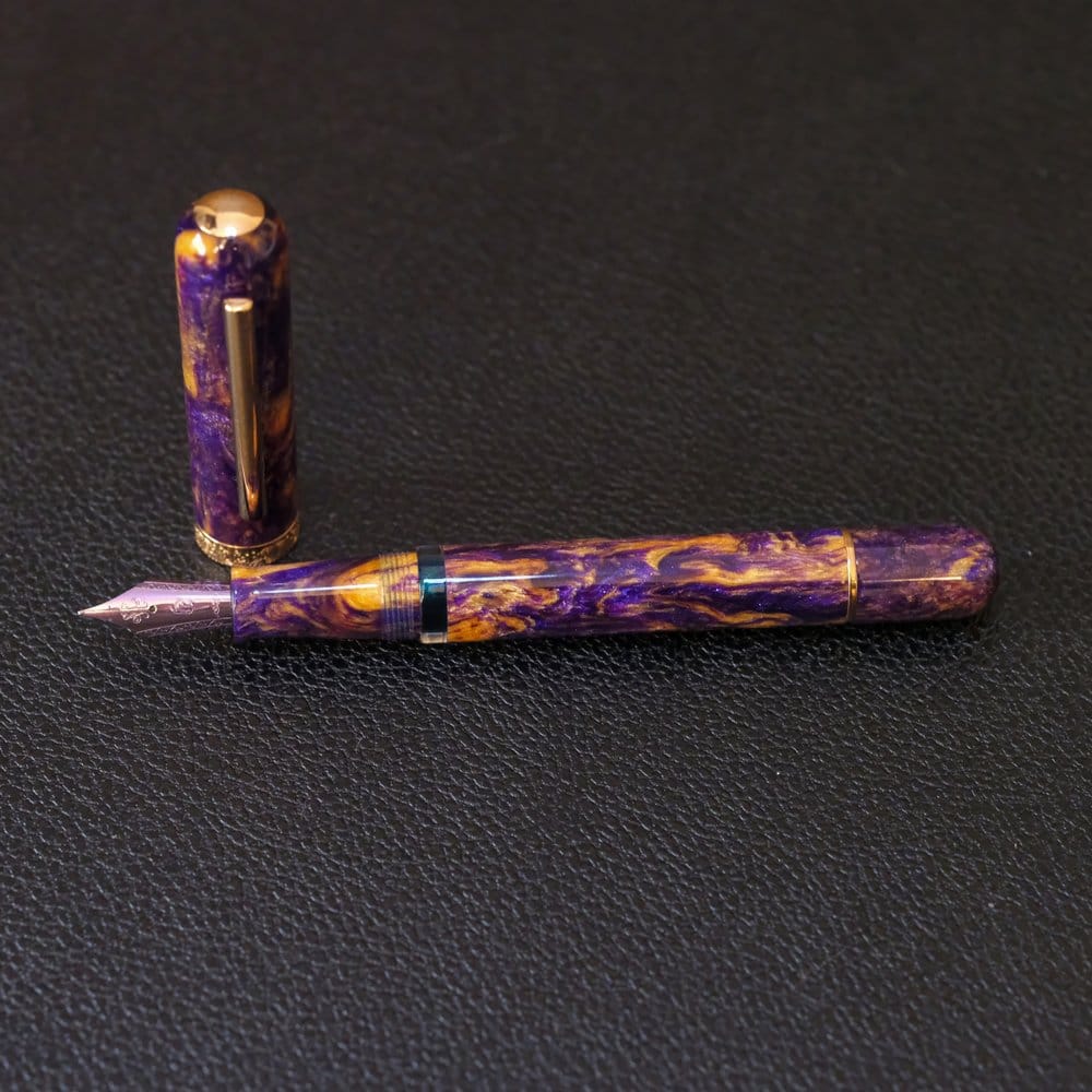 The Nahvalur Voyage, a cylindrical pen with rounded ends, has a purple and gold swirled acrylic body and a purple steel nib, shown against a black, textured background, uncapped