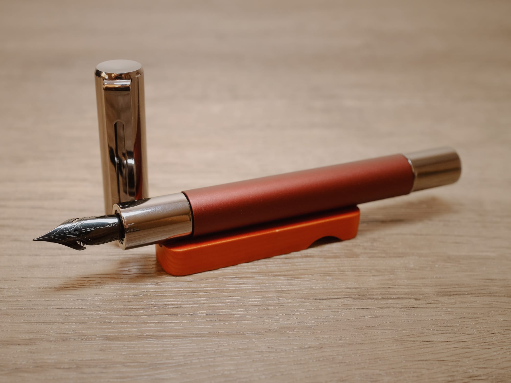 Metal fountain pen with chocolate brown body and gunmetal cap and end, sitting uncapped on a reddish wooden pen rest, showing a dark gray, steel, flex nib