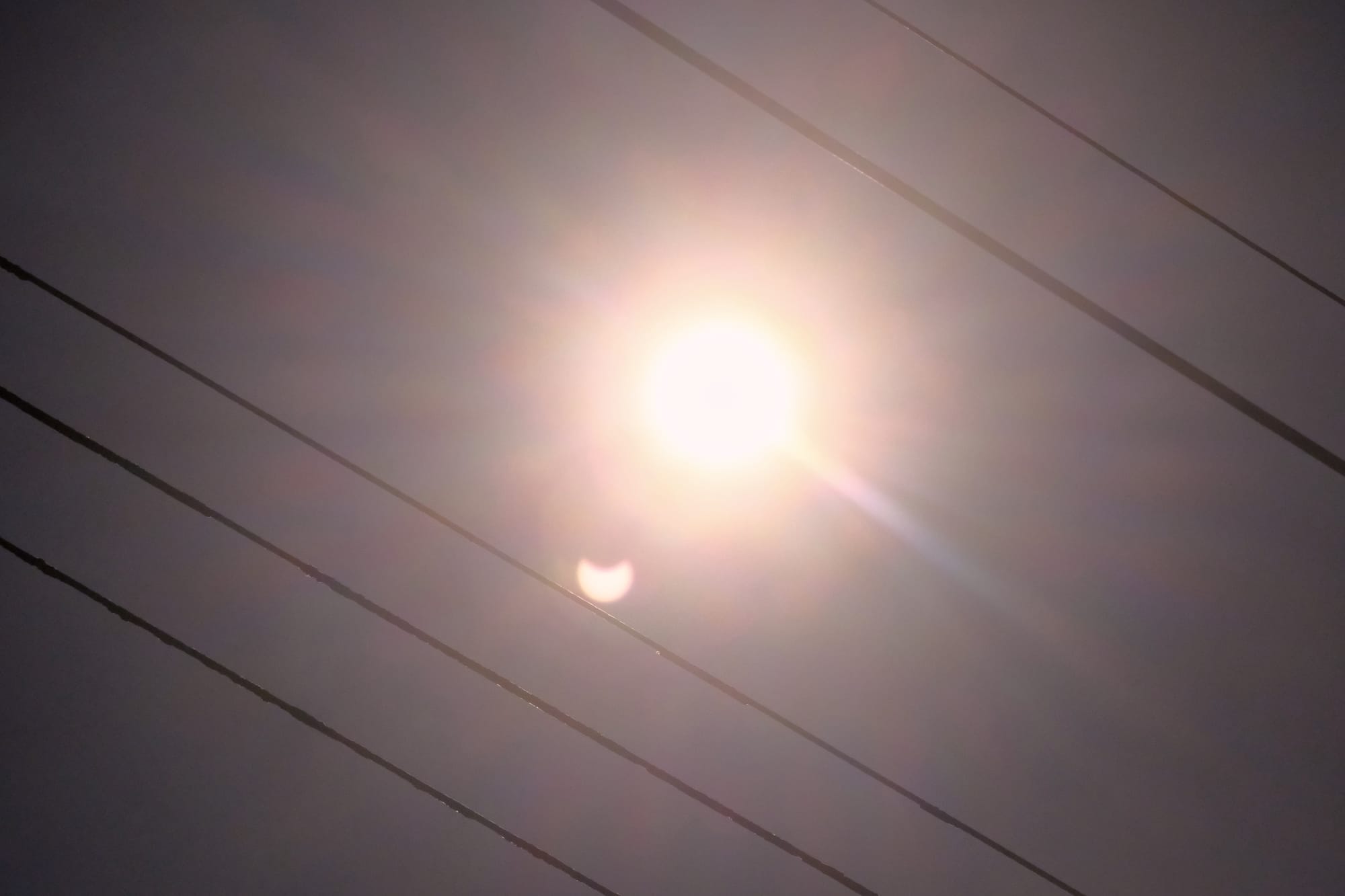 Diagonal power lines framing an overexposed eclipse in the middle of the frame, with a smaller, upside-down, eclipse-shaped reflection below the overexposed sun.