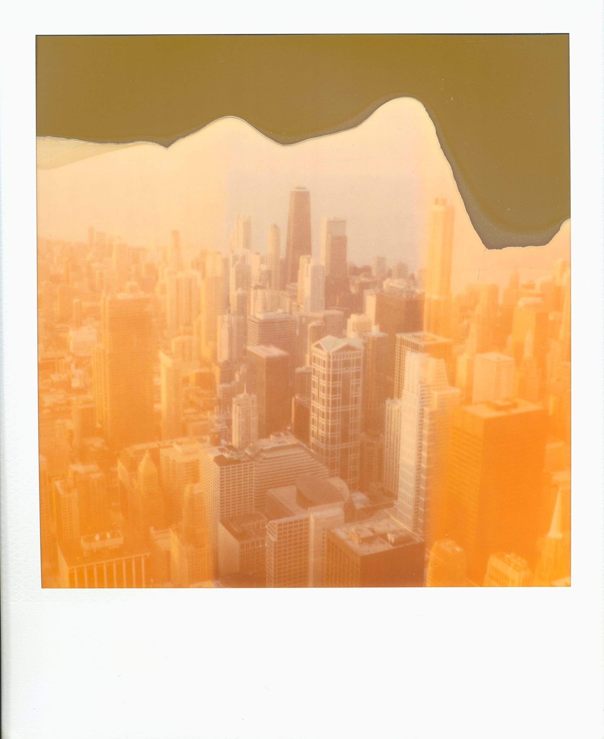 Scan of expired Polaroid film image of the Chicago skyscrapers as seen from the top of the Sears Tower. The image has strongly orange areas on the left and right, and a very desaturated exposure down the middle. The top of the image isn't fully developed, leaving an irregular edge of gray plastic where the sky would be.