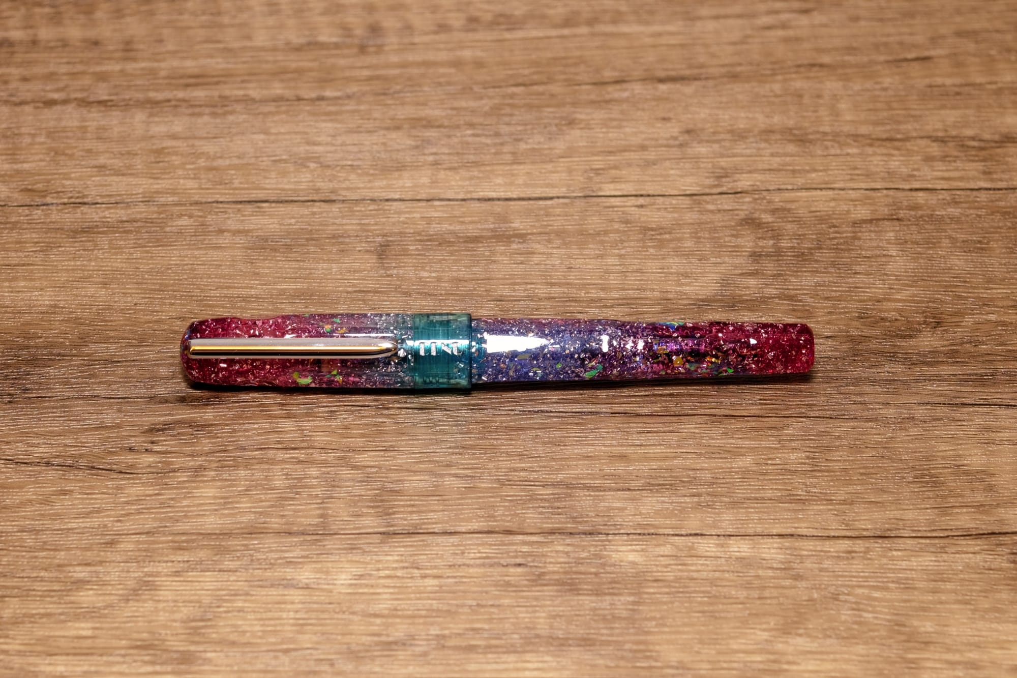 A fountain pen with a dark pink to aqua to lavender to dark pink gradient resin body infused with holographic glitter flakes