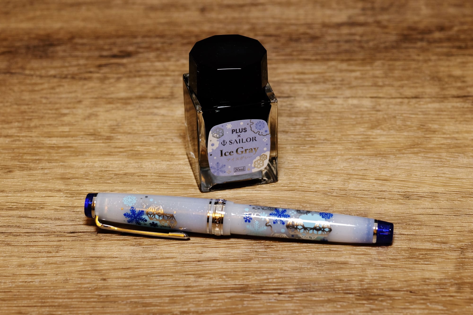 Small glass bottle of fountain pen ink, Ice Gray, sitting behind a fountain pen with a translucent white body with glitter embedded and snowflake graphics in various shades of blue and silver painted on, and dark blue top and bottom