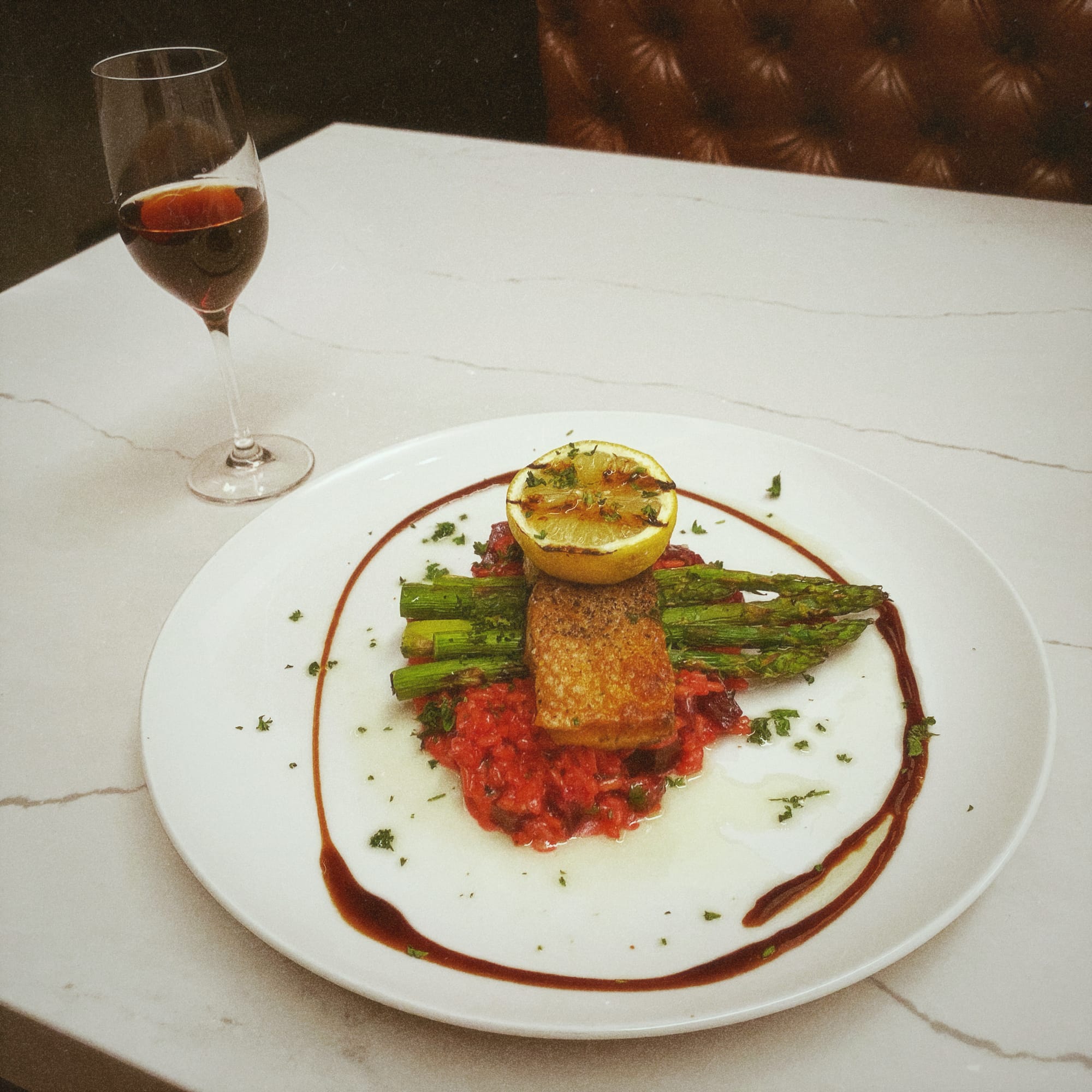 Large white plate with beet risotto piled in the middle, with stalks of asparagus, a filet of salmon, and a grilled lemon wedge stacked sequentially on top. A drizzle of a dark sauce encircles the food.