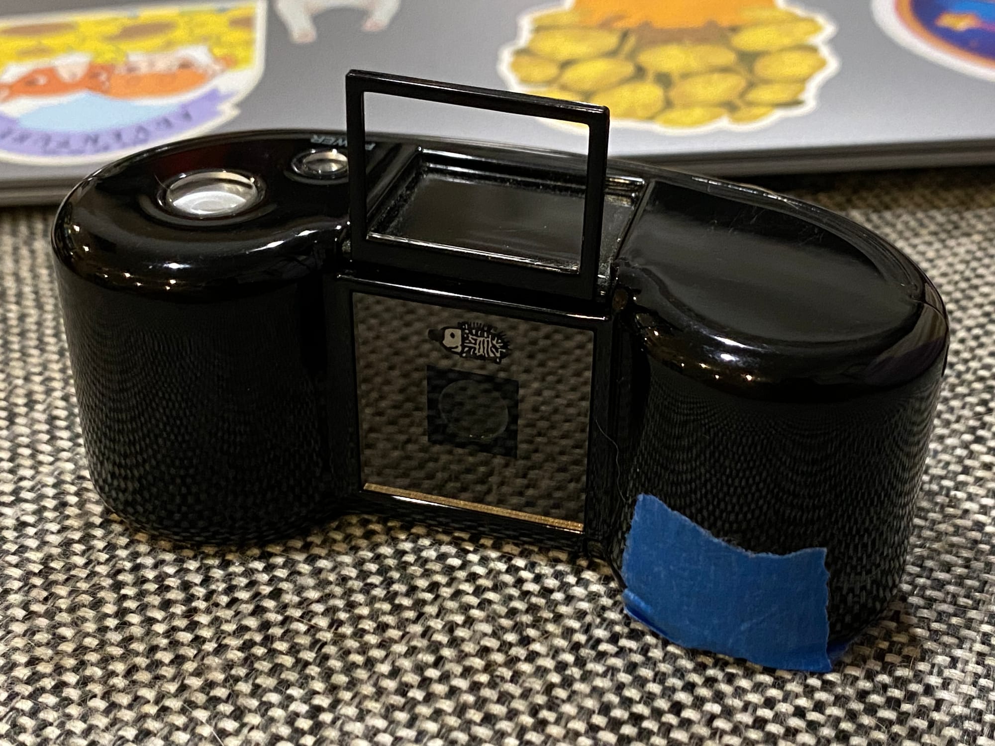 Close up of the black Digital Harinezumi 2 camera, shaped like an oversized roll of 110 film, with a mirrored square around the lens, and the "sport viewfinder" opened up