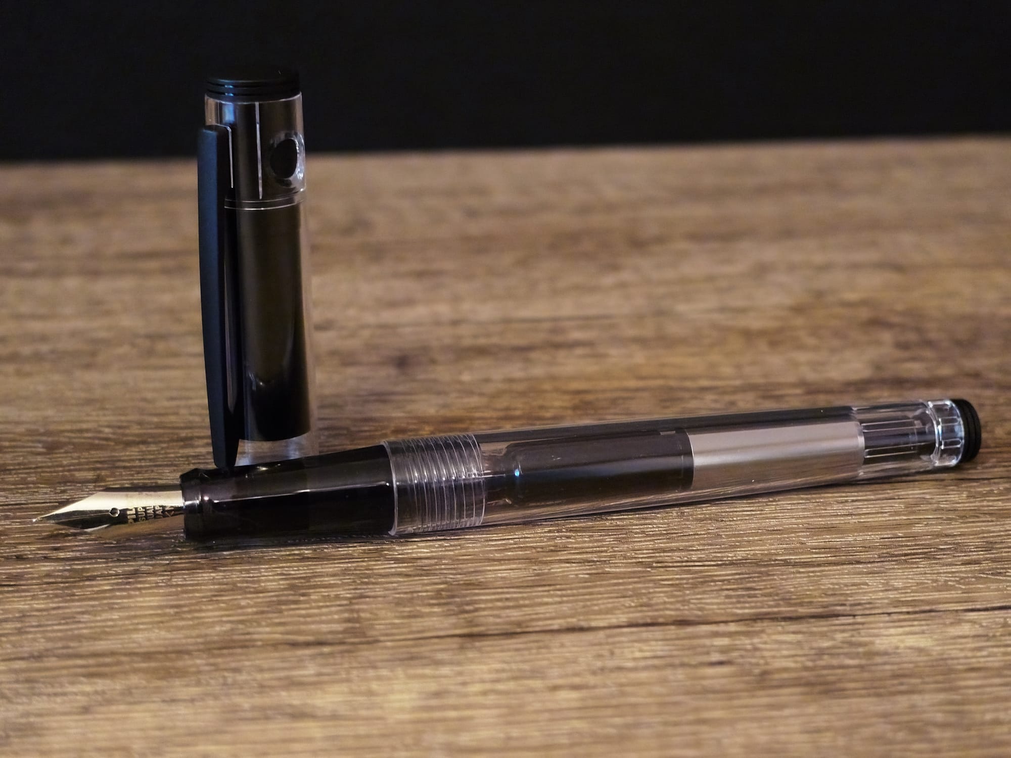 A clear bodied fountain pen uncapped, with its black cap sitting upright behind its black grip section