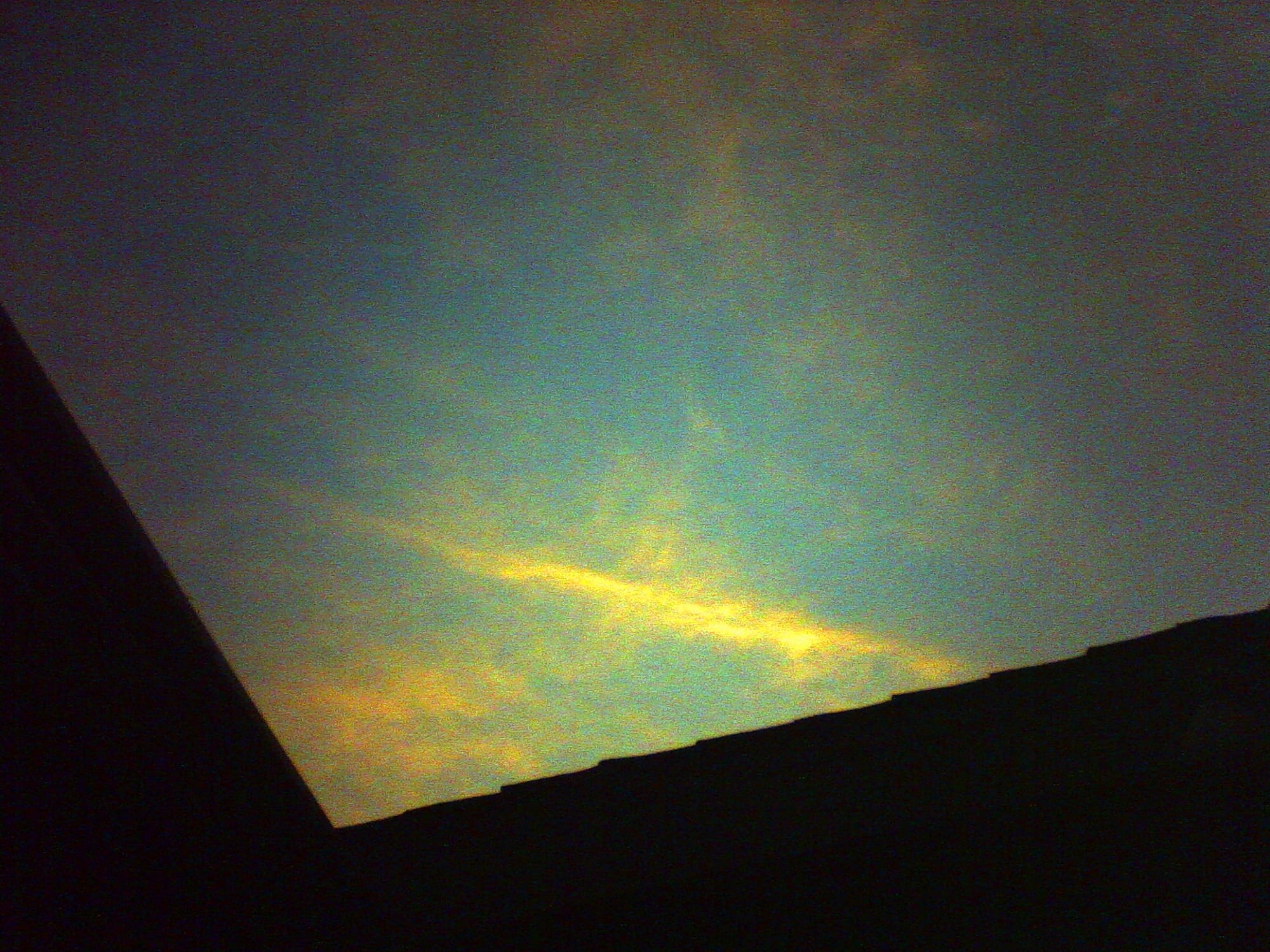 Lo-fi, very grainy shot of overexposed, orange-yellowy, wispy clouds at sunset in a light blue sky with a dark silhouette of a roof framing the bottom of the image