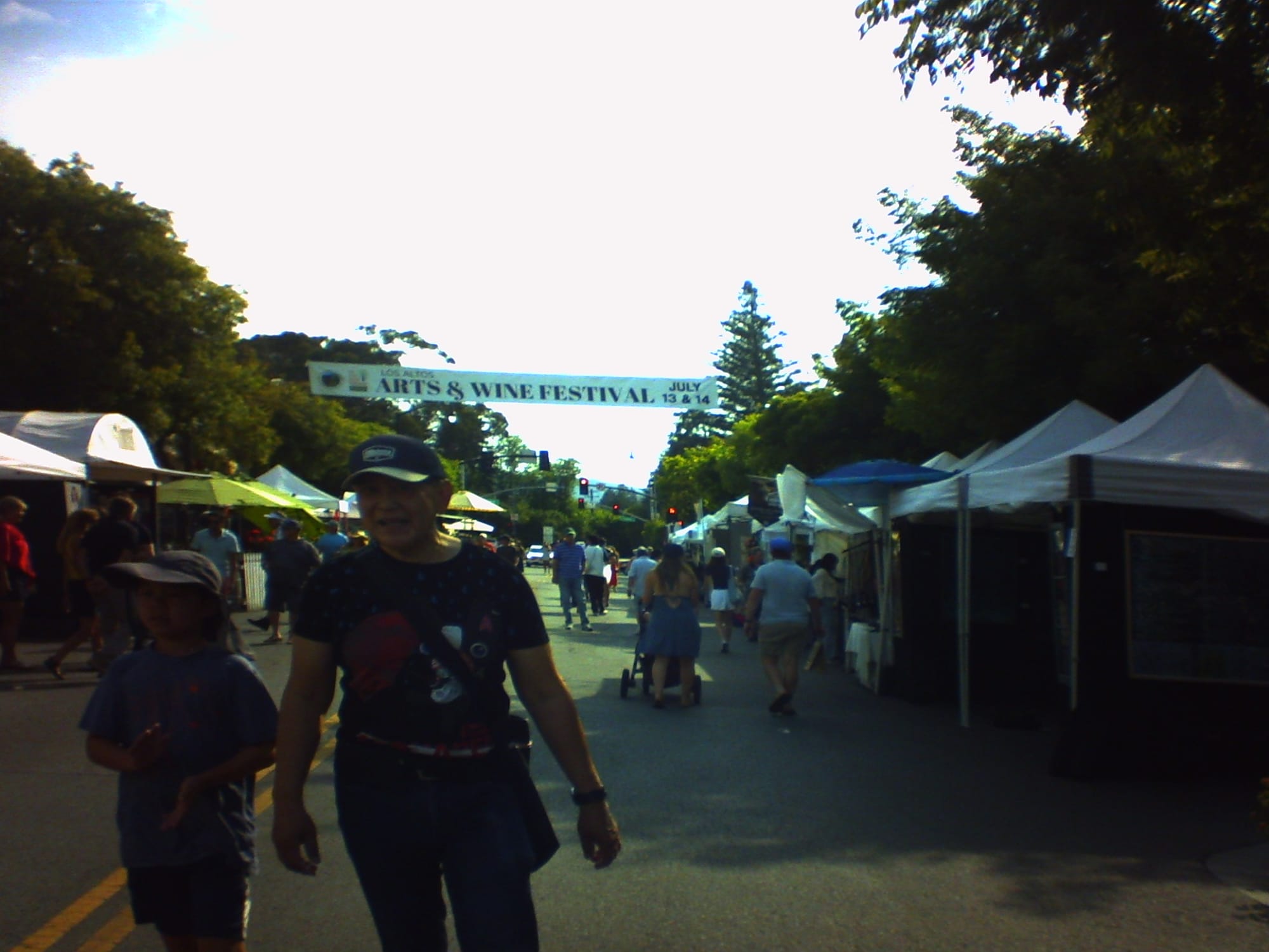 View of a street blocked off for the art & wine festival, lined with vendor tents on either side, with a banner, "Los Altos Arts & Wine Festival, July 13 & 14" strung above it. A couple people in the foreground are slightly "wobbly" due to a rolling shutter effect.