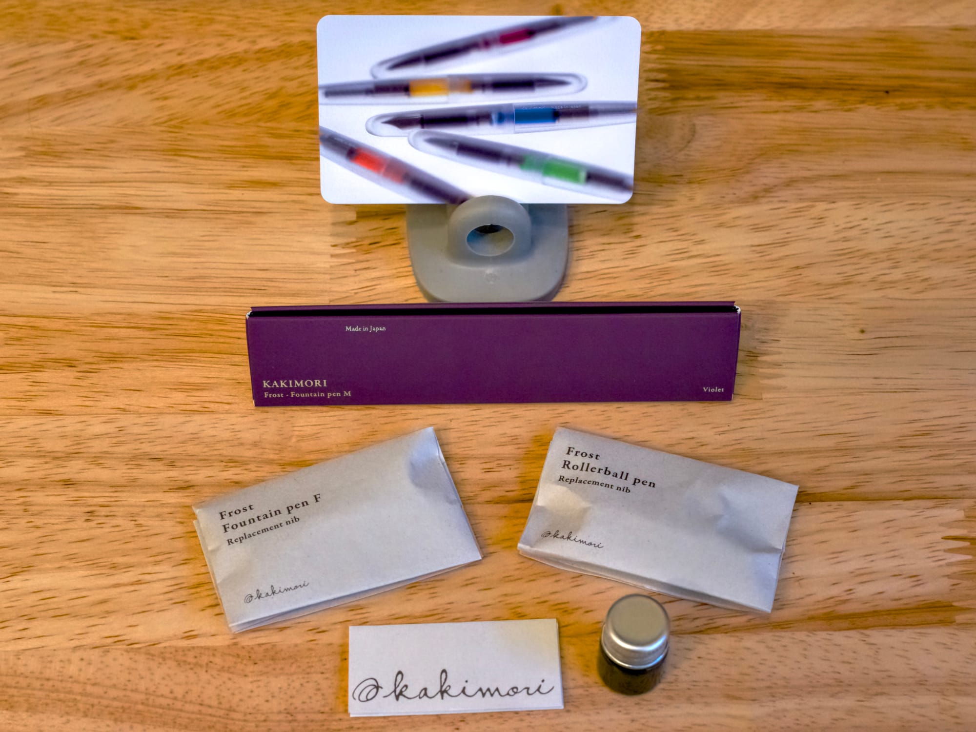 Purple Toblerone-shaped cardboard package with 2 replacement nibs in folded paper packaging, a decorative card showing various clear translucent pens behind the pen box, and a small glass bottle of ink in the very front of the group with a small leaflet