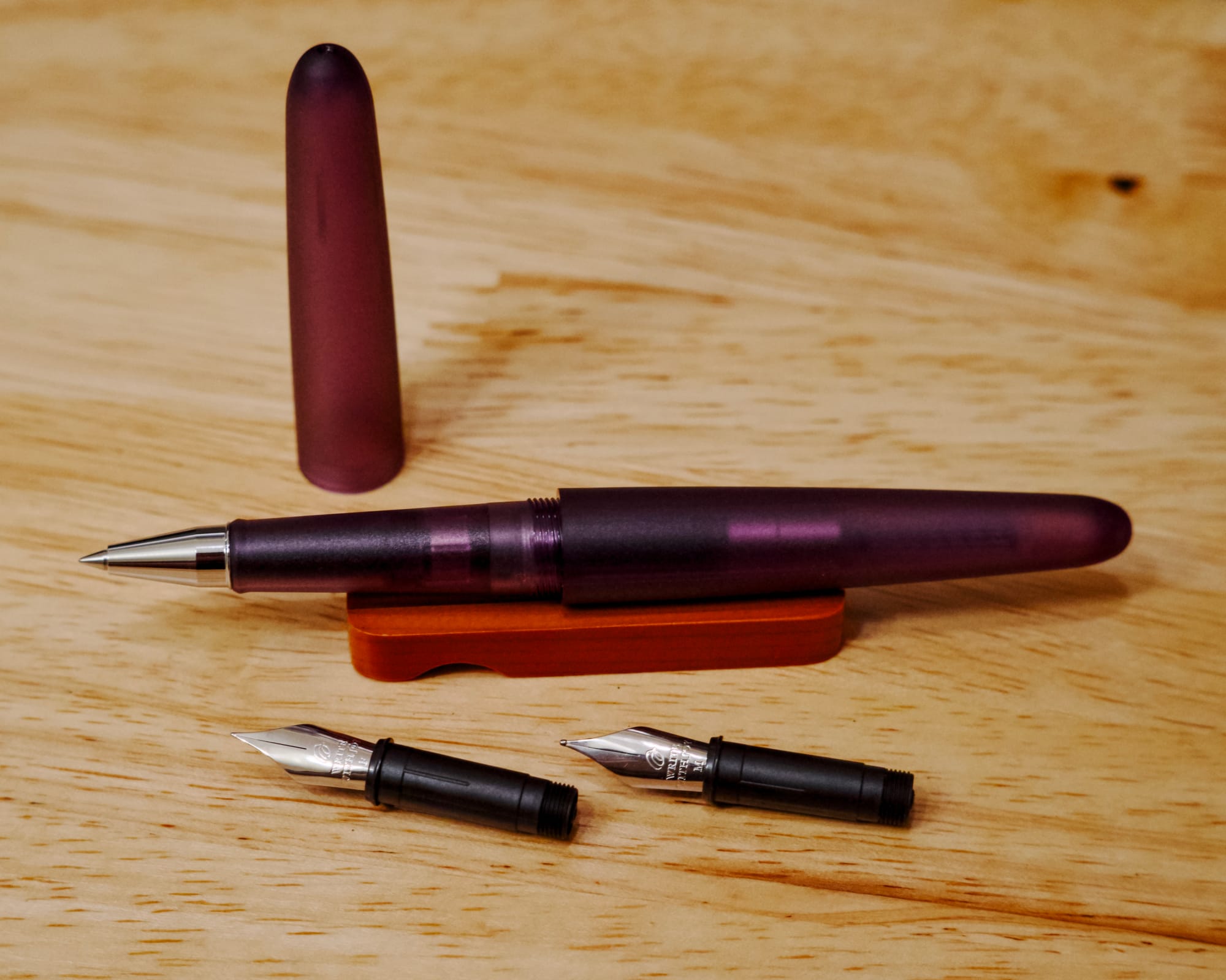 Translucent purple rollerball pen sitting on orange wooden pen stand with 2 nib units sitting on desk in front, plastic cap behind