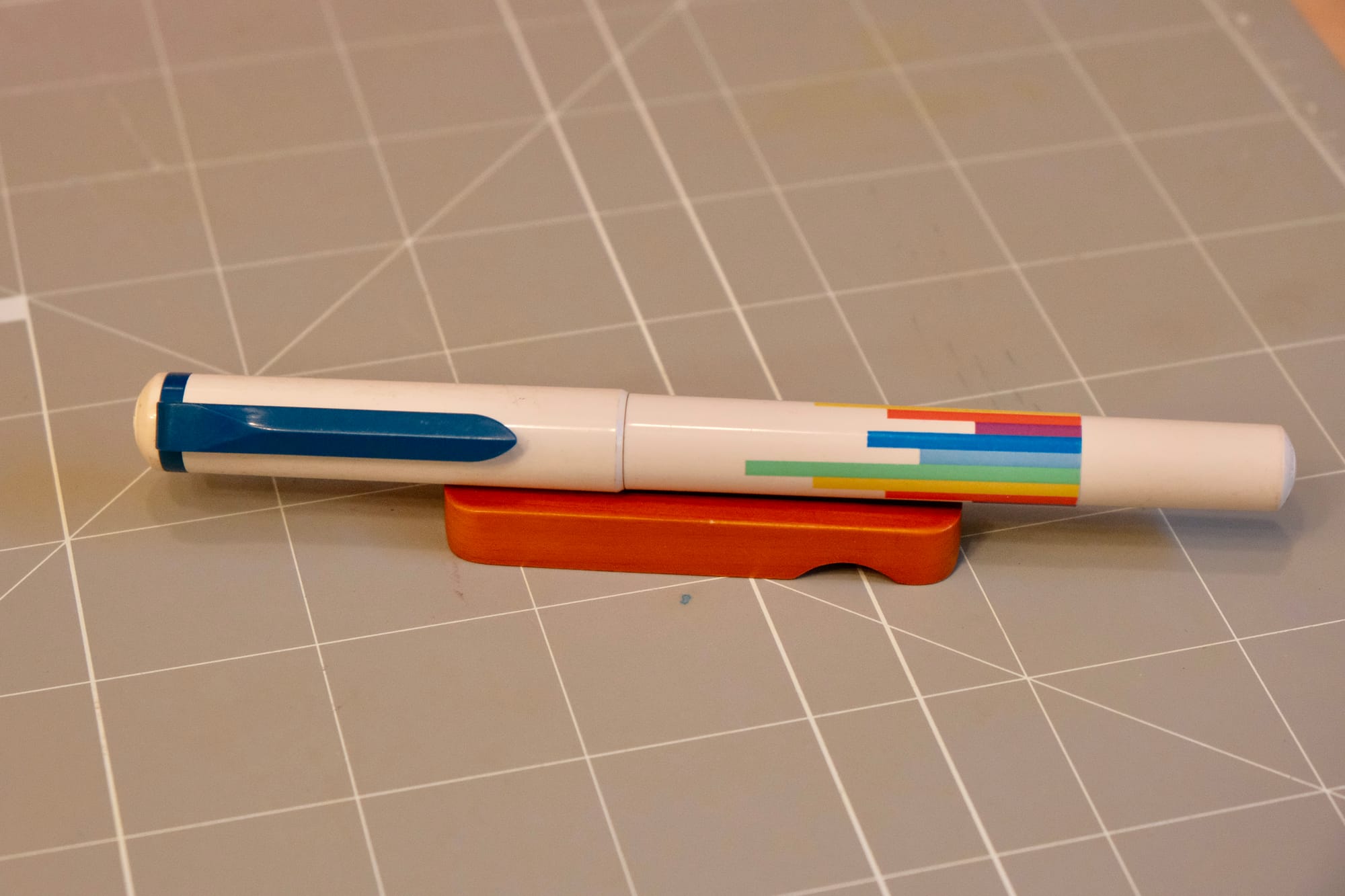 A white plastic pen with a dark blue clip and a "bar graph" design of vertical bars with varying lengths in range of rainbow colors on the pen body