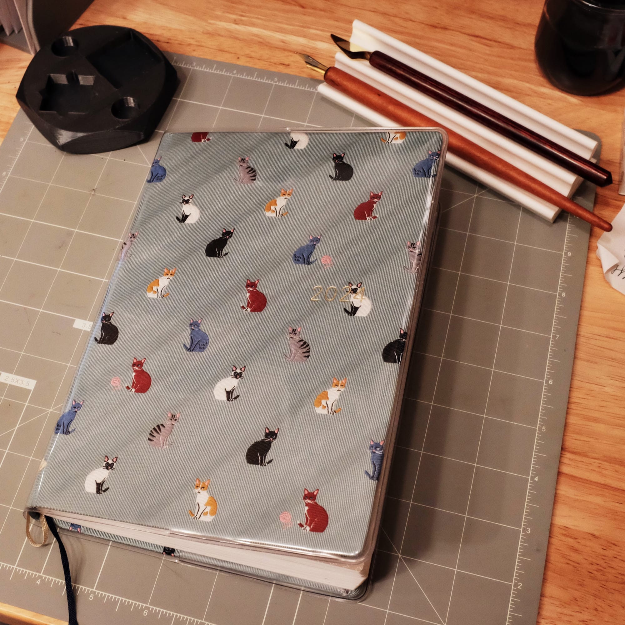 A notebook with a fabric cover showing various colored cats in a grid arrangement. The notebook is on a gray cutting mat on a wooden desk with a 3D printed ink bottle holder on the top left, and a 2-pen holder on the top right holding a couple dip pens