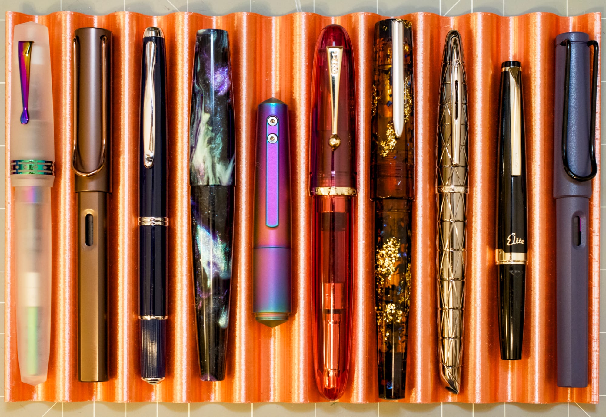 A copper-colored 3D-printed pen tray holding 10 different fountain pens with varying sizes, shapes, designs, and colors