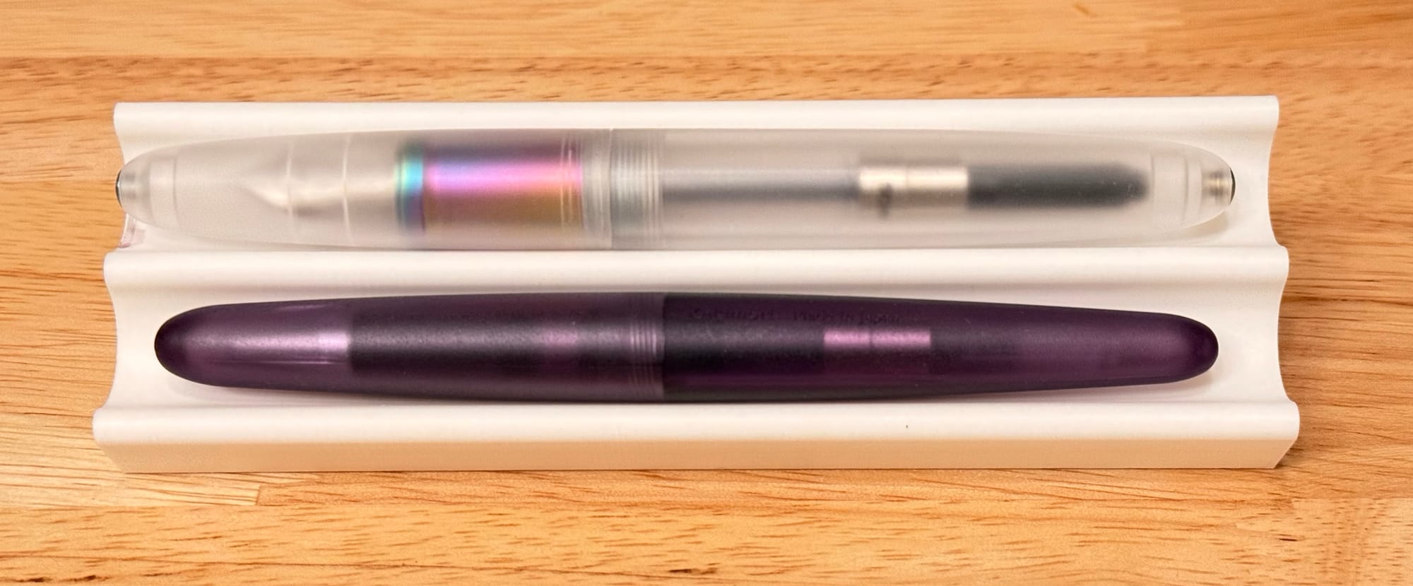 White dual pen stand with one frosted translucent fountain pen in the top slot with a rainbow anodized grip section visible through the cap, and a purple frosted rollerball pen with a similar cigar-shape in the bottom slot