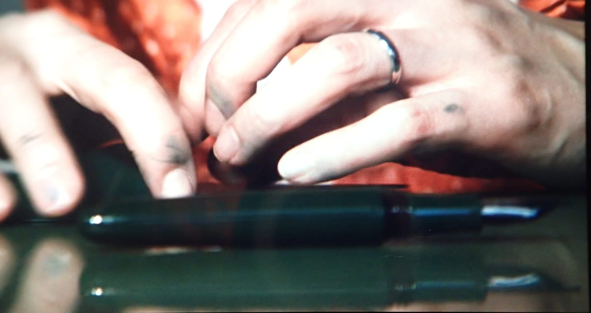 Zoomed in on a green (lacquered?) fountain pen sitting on a reflective table top and ink-stained hands in the background