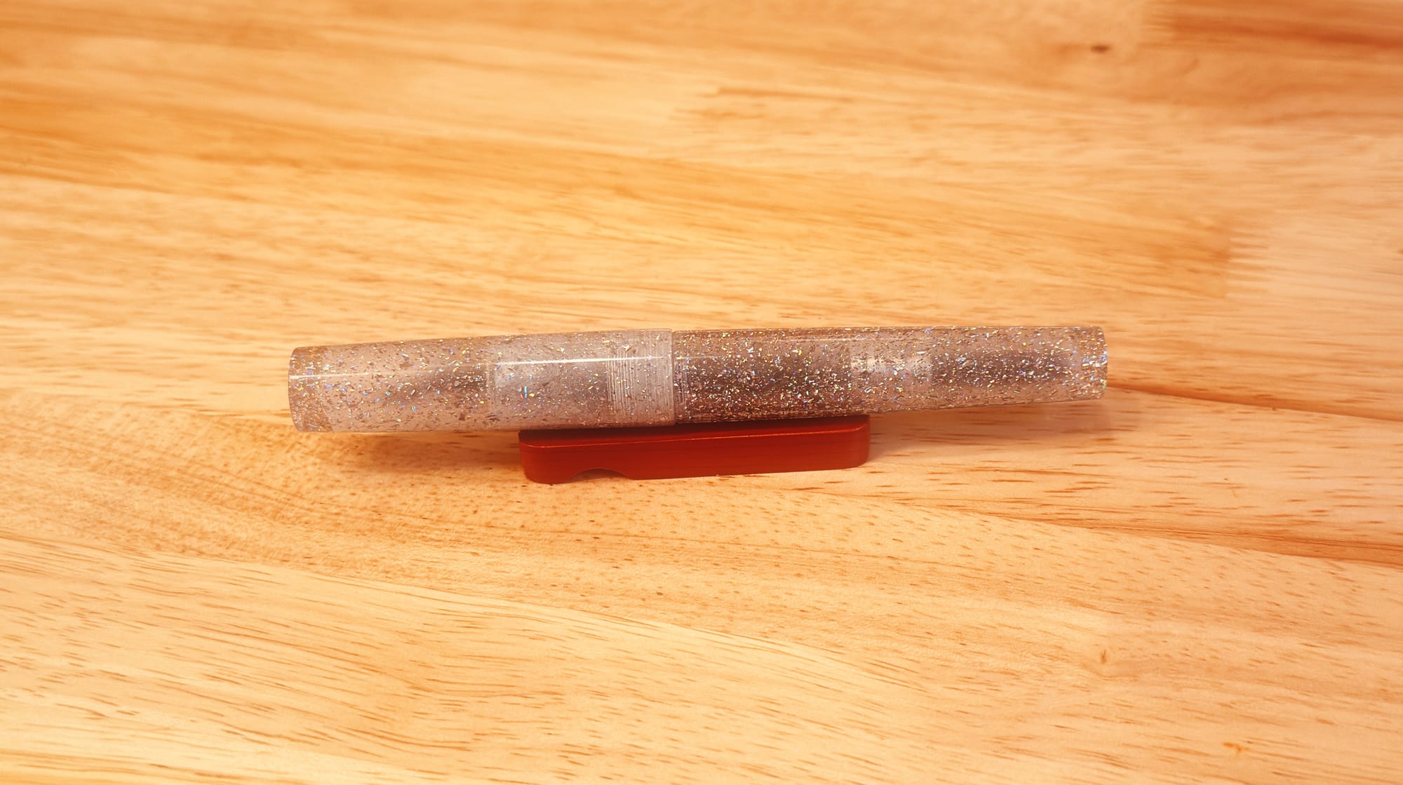 A fountain pen sitting on a small wooden pen rest: it has translucent clear resin packed with a ton of silver holographic glitter.