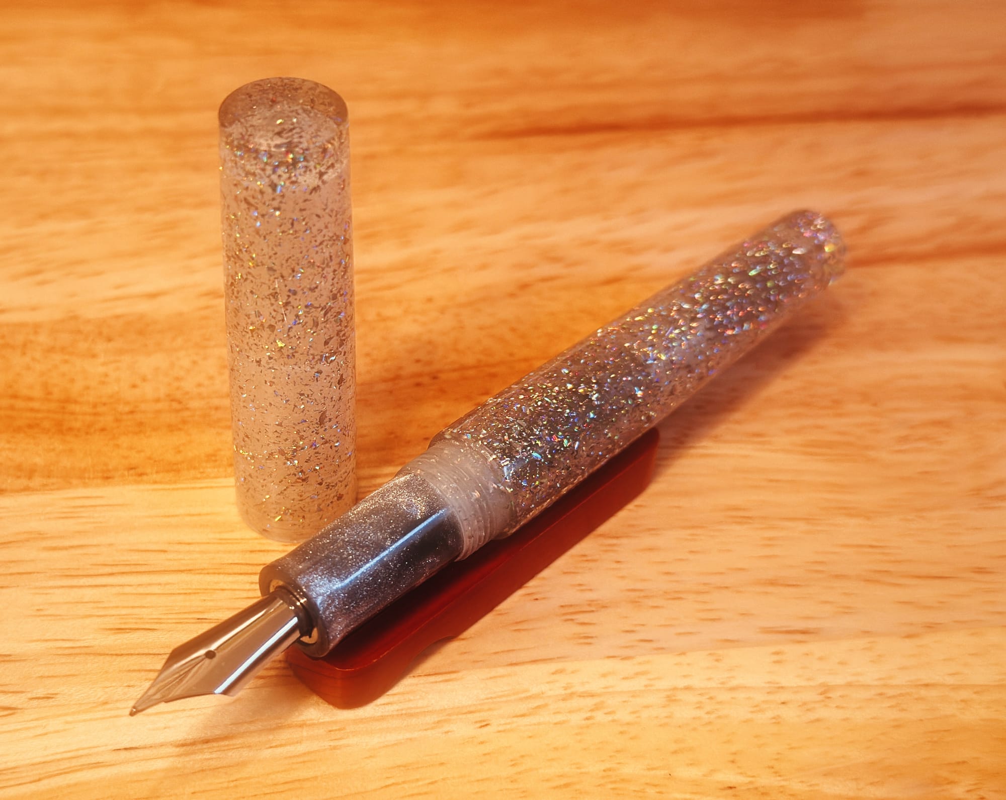 A fountain pen sitting on a small wooden pen rest, uncapped, it's cap sitting vertically behind it. It has translucent clear resin packed with a ton of silver holographic glitter. Its grip section is a slightly different resin that is medium gray, shot through with dense silver microglitter. Its nib is silver, with a custom ground tip.