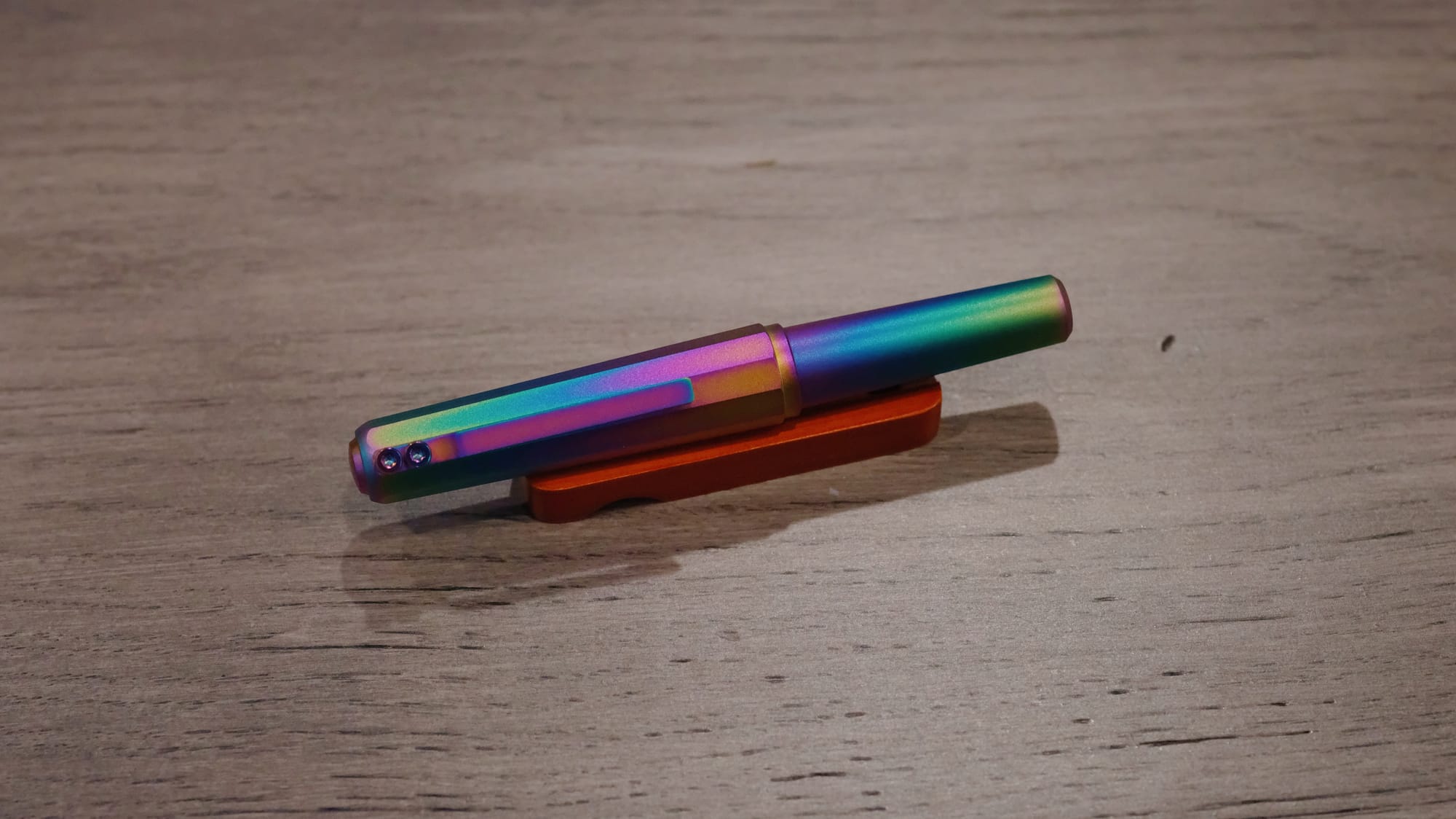 Rainbow anodized metal pen sitting on a small wooden pen rest on a wooden surface