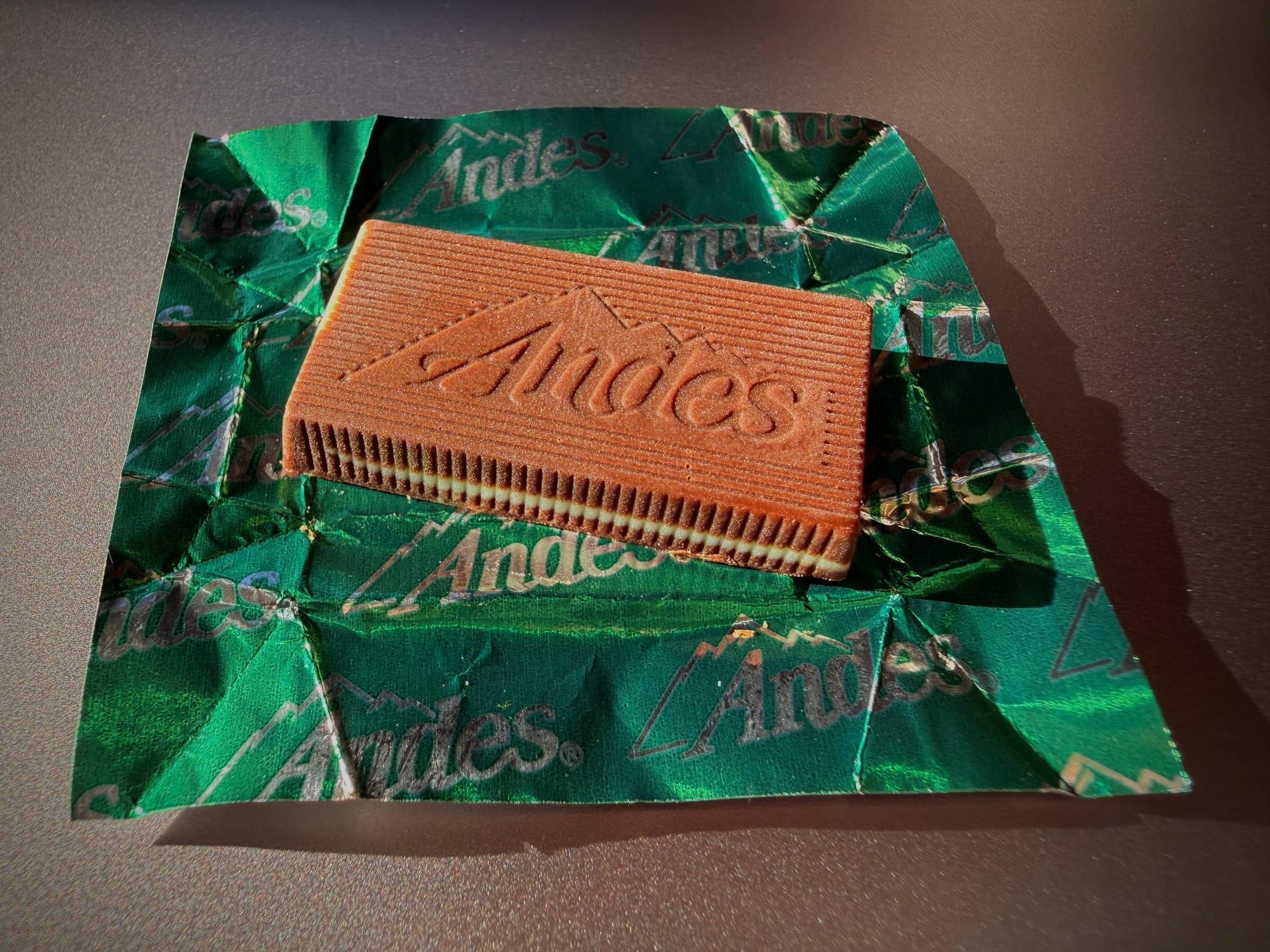 Close up of an Andes mint chocolate candy sitting on top of an open Andes wrapper, showing off its shiny green color and silver Andes logos