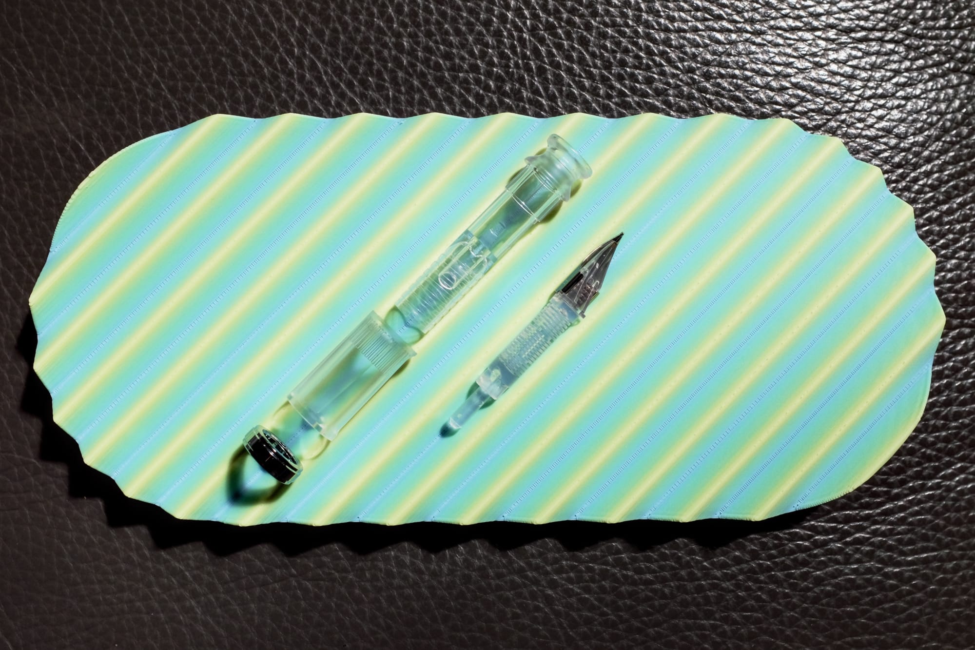 A 3D-printed pen tray with diagonal ridges and a gradient color scheme from light blue at lowest layers, to a gradually lightening pastel green, and yellow at the tops of the ridges; clear fountain pen section parts disassembled and placed on the tray with the section parts together on one slot, and the feed and nib in another