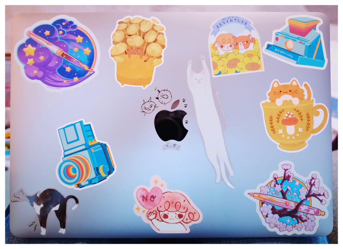 MacBook Air lid showing various stickers of stylized fountain pens, cats, vintage cameras, and Pepper by Lovesoup