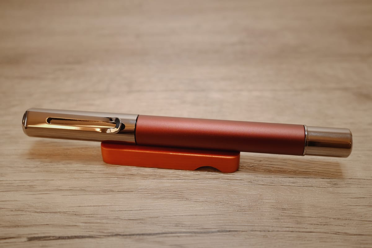 A metal pen with a chocolate brown body and gunmetal cap and end, sitting on a reddish wooden pen rest