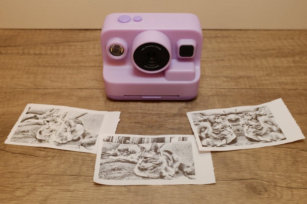 A purple plastic camera with a toy-like appearance and 3 grayscale thermal prints from it laid out in the foreground
