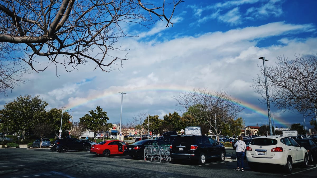 A wide view of a rainbow in a blue, cloudy sky as seen from a parking lot with several cars in the foreground