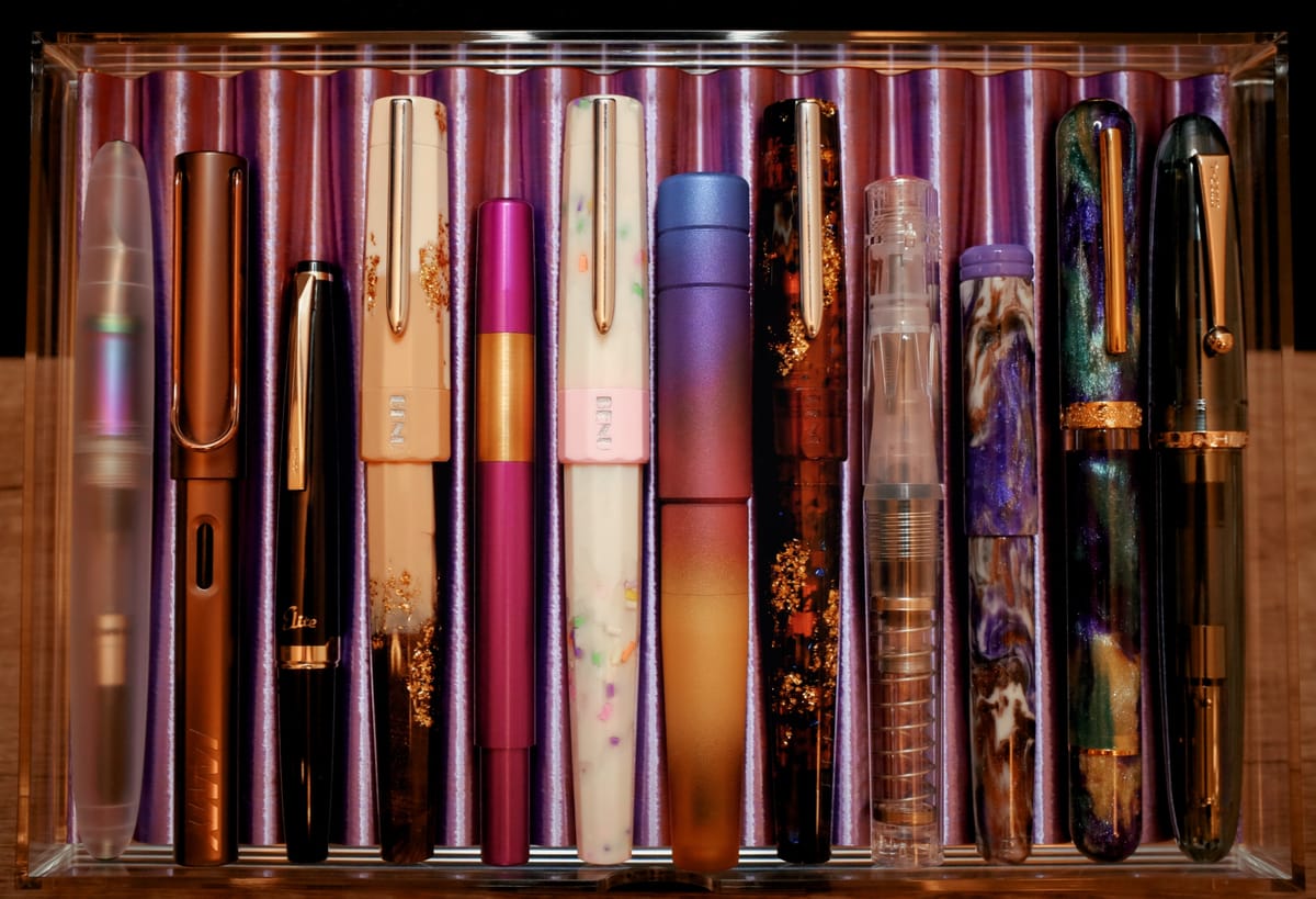 An acrylic drawer with a purple 3D-printed tray holding 12 various fountain pens of different designs and sizes