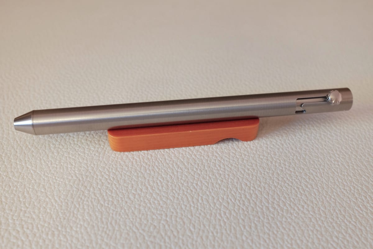 Minimal metal fountain pen with a bolt-action mechanism sitting on a small wooden pen rest on top of a white textured surface