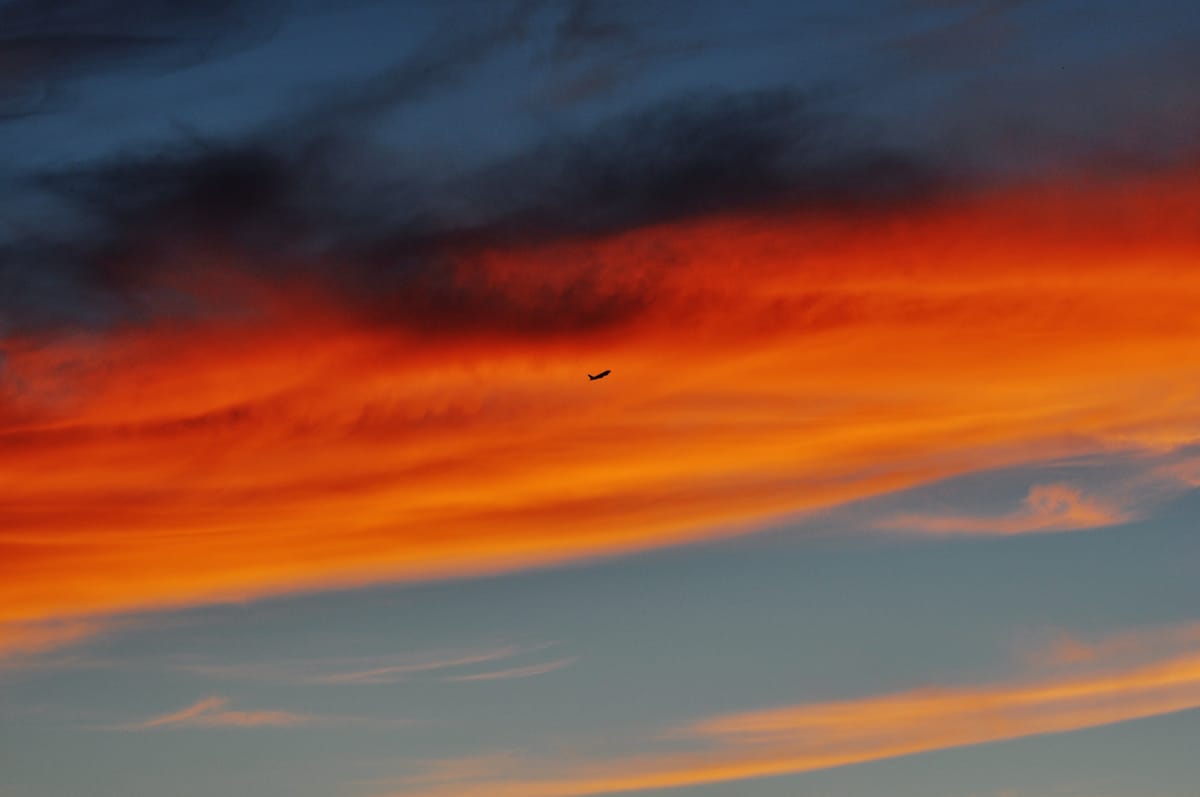 Telephoto image of a sunset sky with blazing red-orange clouds and a tiny silhouette of an airplane in front