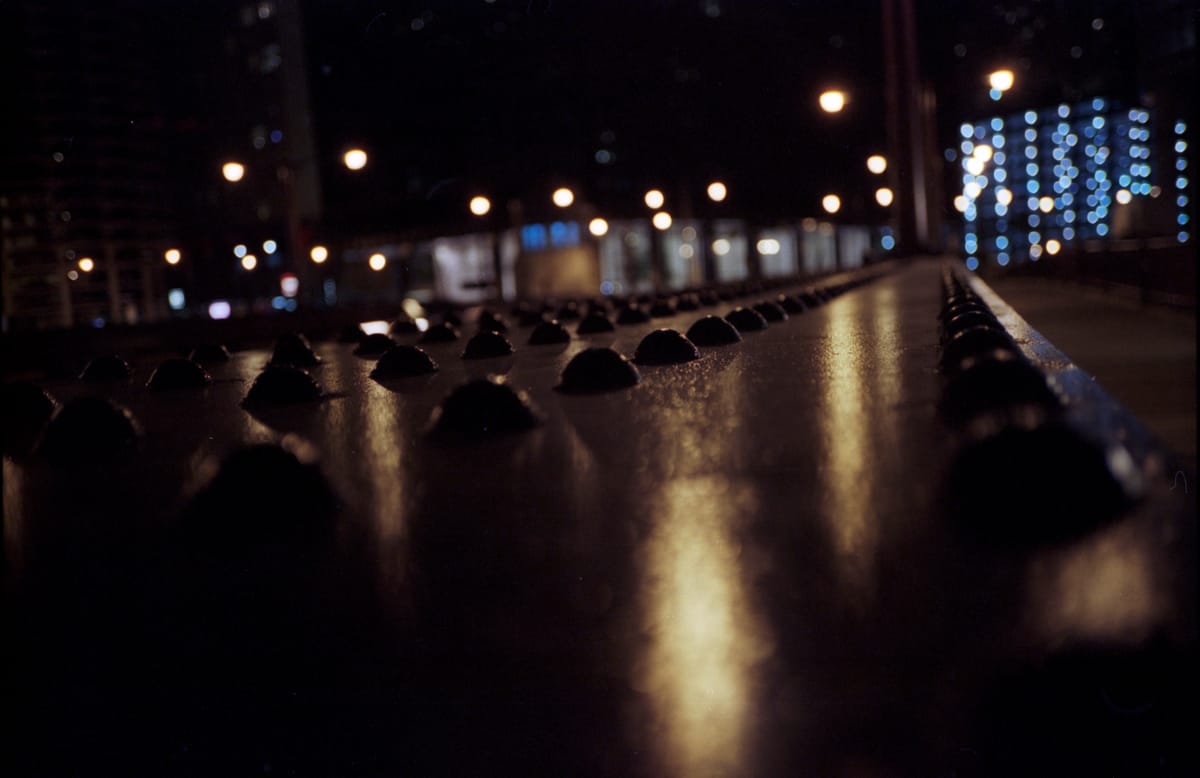 Night time picture of rivets on a Chicago bridge's railing by the sidewalk with out-of-focus lights in the background