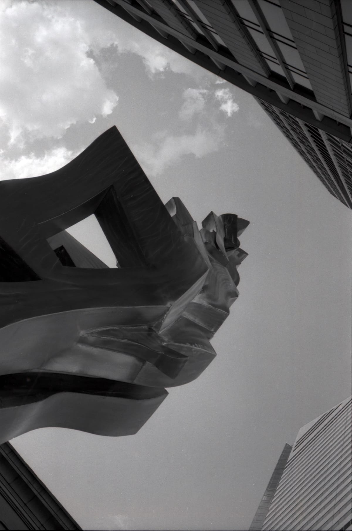 Looking up at an abstract, undulating metal sculpture amidst skyscrapers in Chicago, some poofy clouds in top left