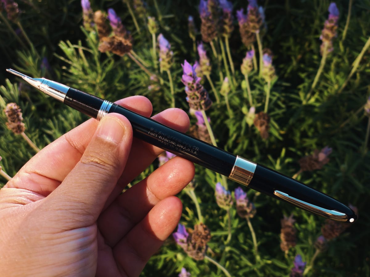 A hand holding Sheaffer Snorkel fountain pen in front of a lavender bush with its "snorkel" extended beyond the nib