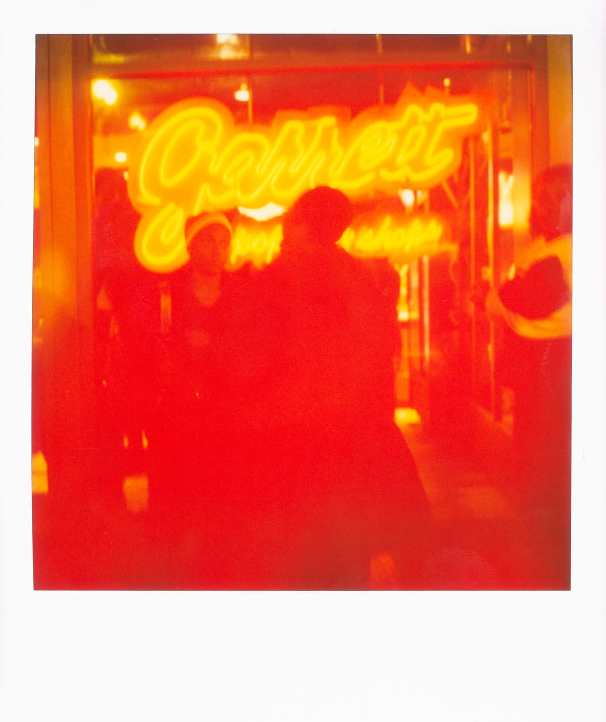 Scan of a Polaroid picture of a couple people standing in front of a bright red neon sign, "Garrett"