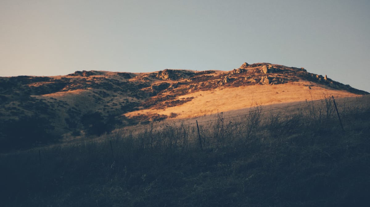 Hills by the roadside in early evening sunlight, some areas covered with scrub, others with large rocks