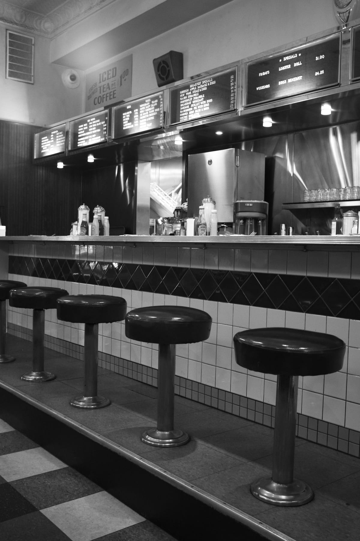 Black and white picture of a counter at a diner with row of short stools on the floor in front, shiny appliances behind