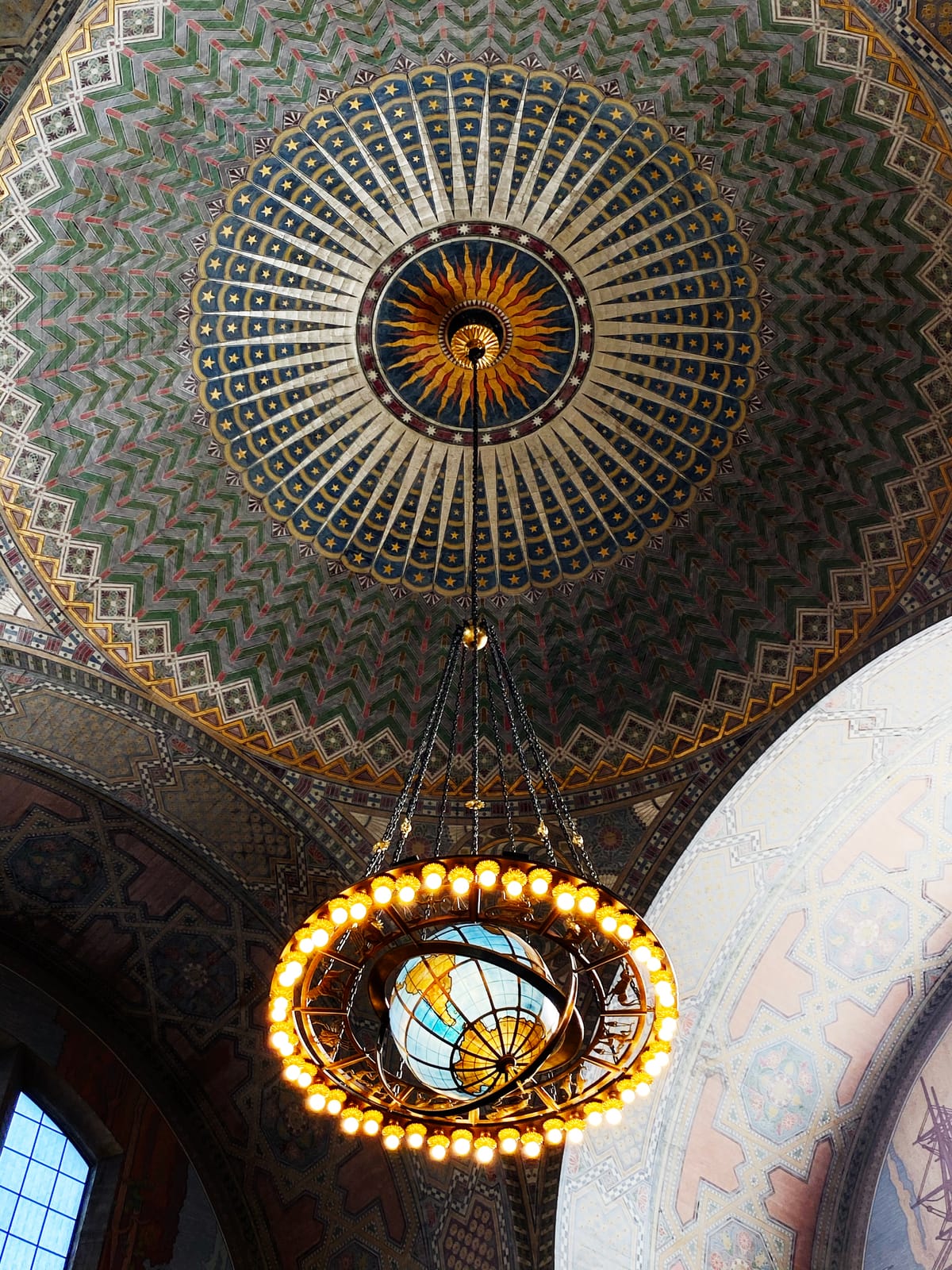 Close up detail of an ornately painted ceiling with a sun design from which a chandelier with a globe center is hanging