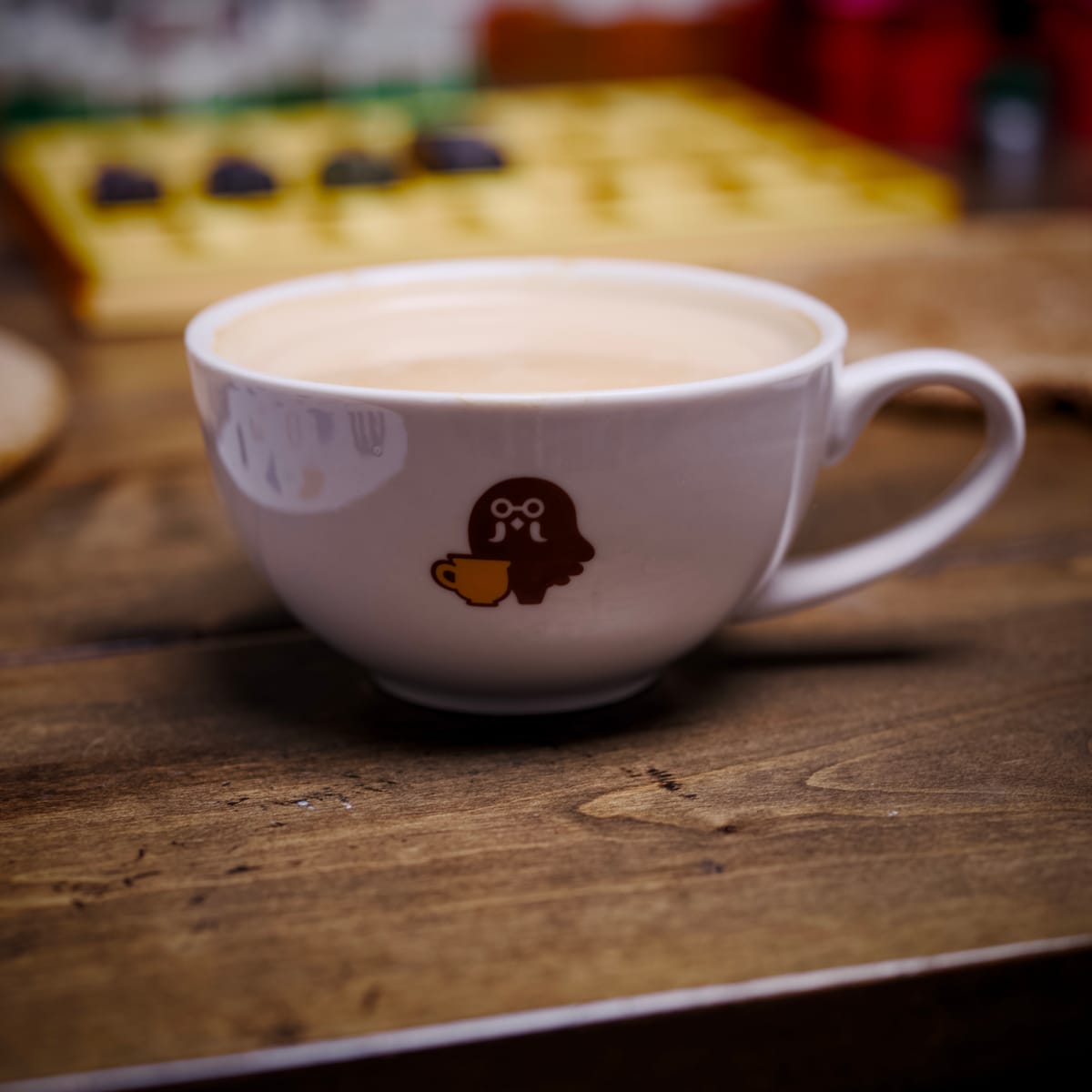 Close up picture of a coffee cup on wooden table with a logo of a stylized pigeon with glasses and a yellow coffee cup