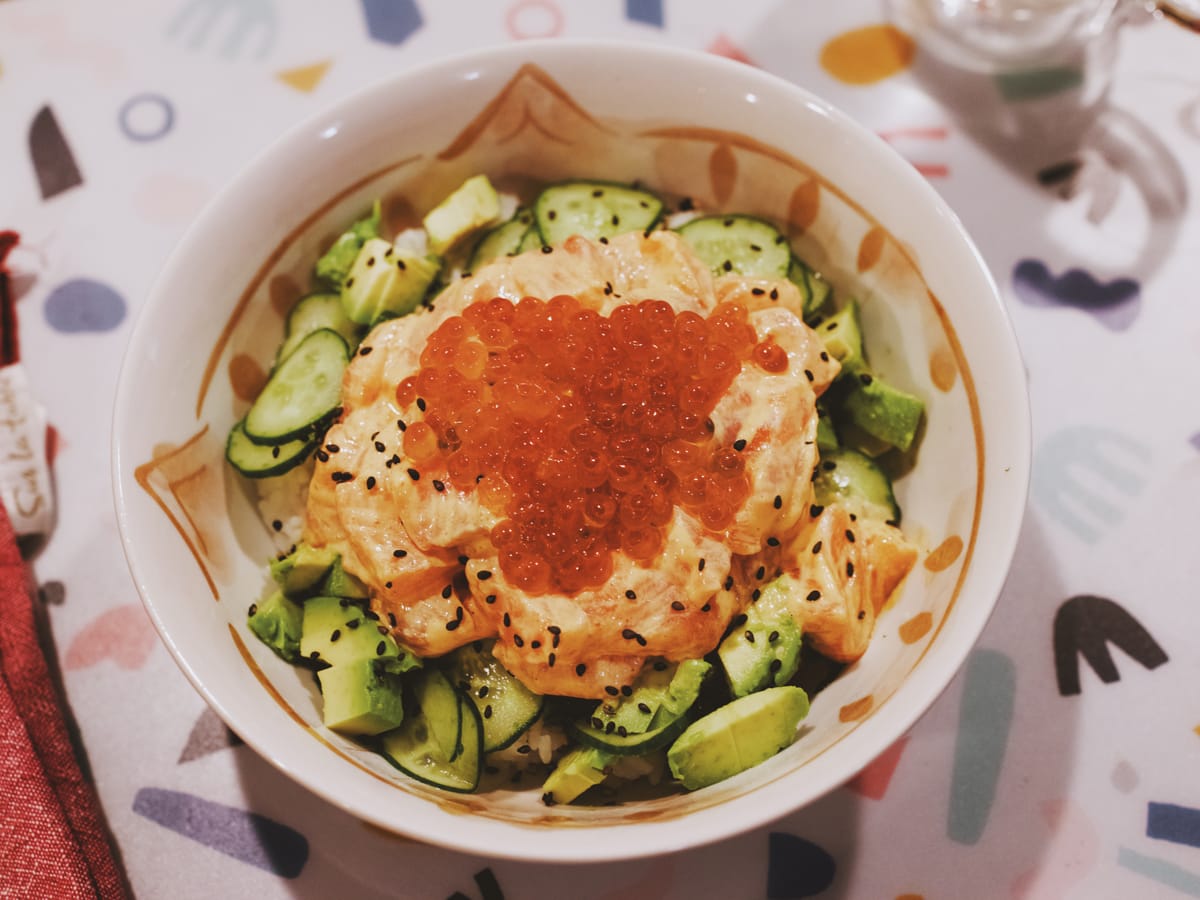 Bowl of rice, cucumber slices, avocado cubes, chunks of salmon with spicy mayo sauce, salmon roe, black sesame seeds on top