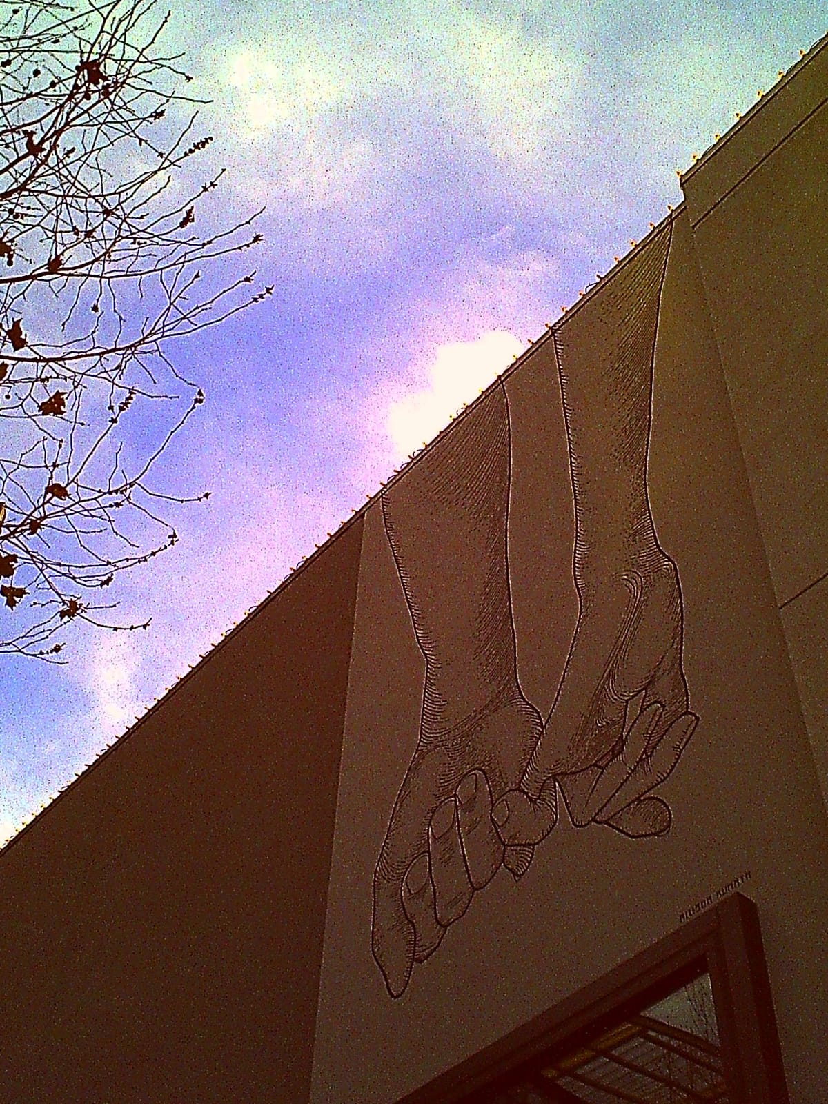 Looking up at side of building with line art of a hand holding the extended pinky of another hand, sky in background