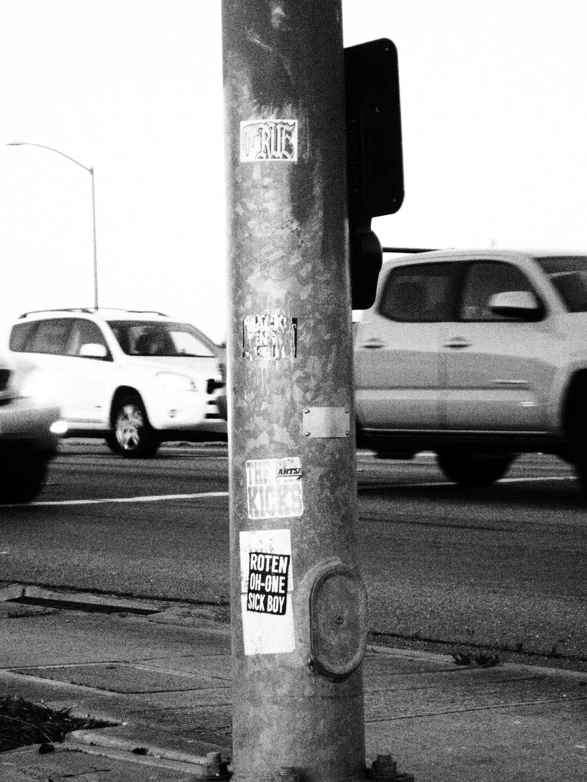 Grainy black and white image of a traffic light pole with random stickers on it, traffic passing behind it