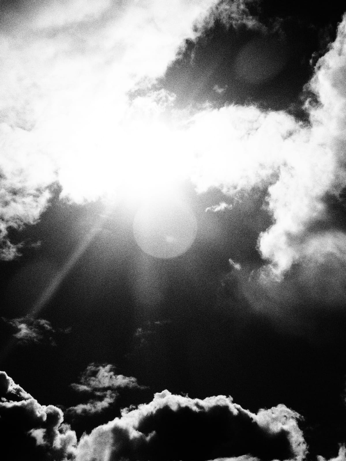 Grainy black and white photo of the sky with poofy clouds on top and bottom of frame and strong sun flare in middle