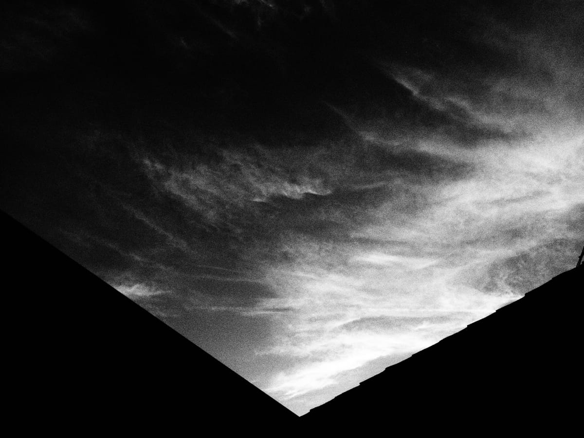 High-contrast black and white image of wispy clouds in the sky with a v-shaped roof line in foreground