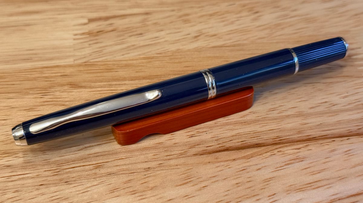 Dark blue capless fountain pen with silver trim sitting on a small wooden pen rest on top of a lighter wooden table