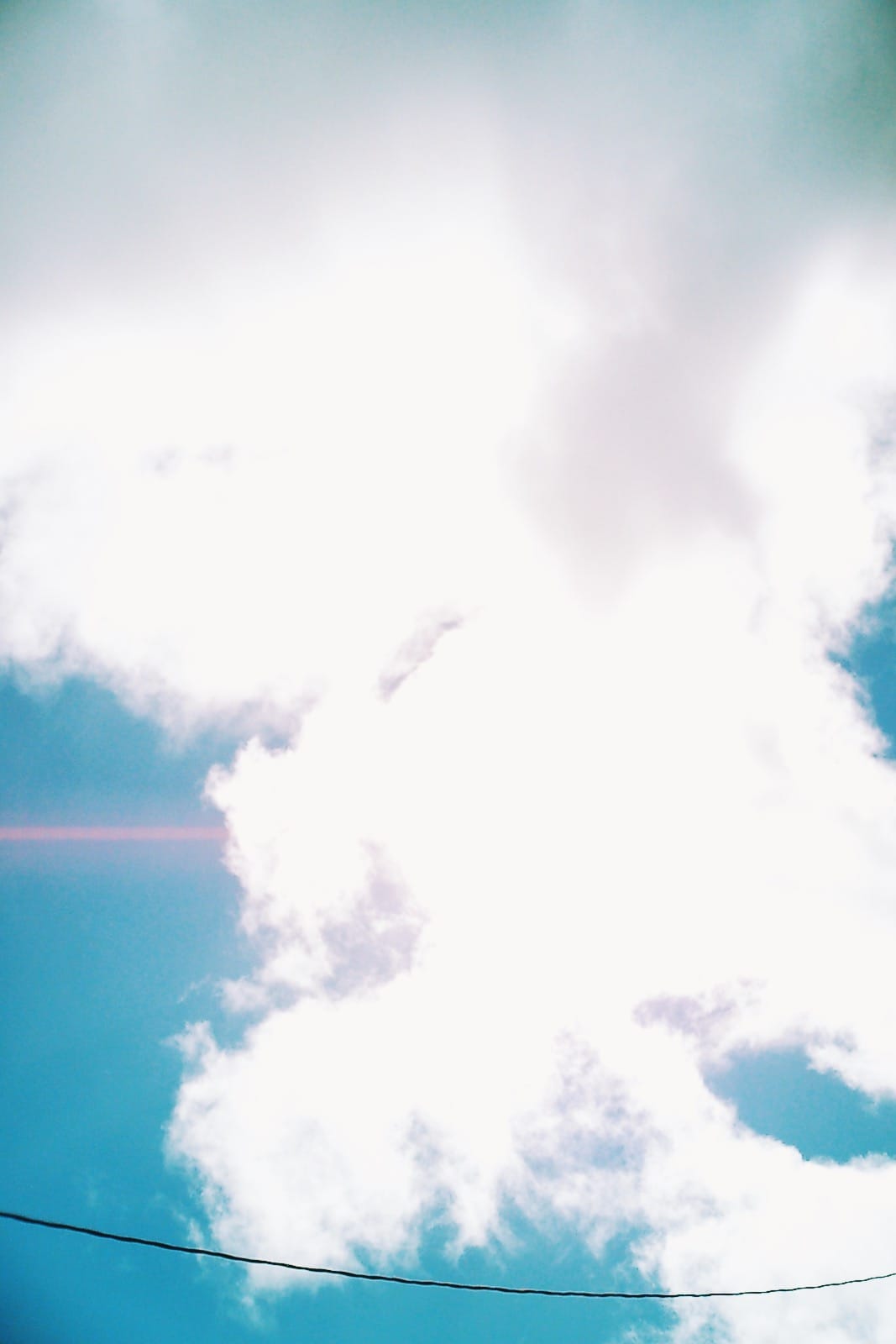 Looking up at a blue sky with poofy white clouds illuminated by a bright afternoon sun, a pink sun flare and power line visible