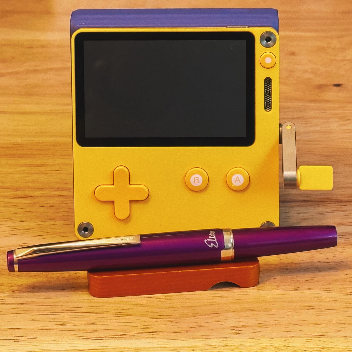 A bright, sunny yellow PlayDate handheld console with its crank extended and purple case, purple Pilot Elite fountain pen resting in front